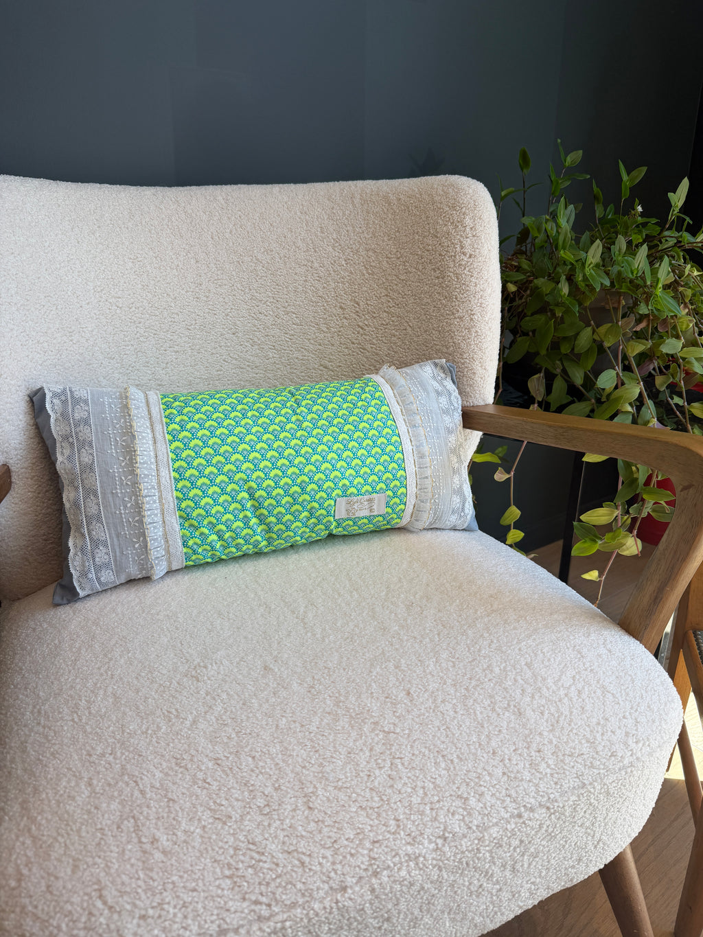 Coussin long fait main Lyli Créarti, tissu imprimé écailles vert et bleu, bandes latérales en dentelle grise, posé sur fauteuil en bouclette blanche