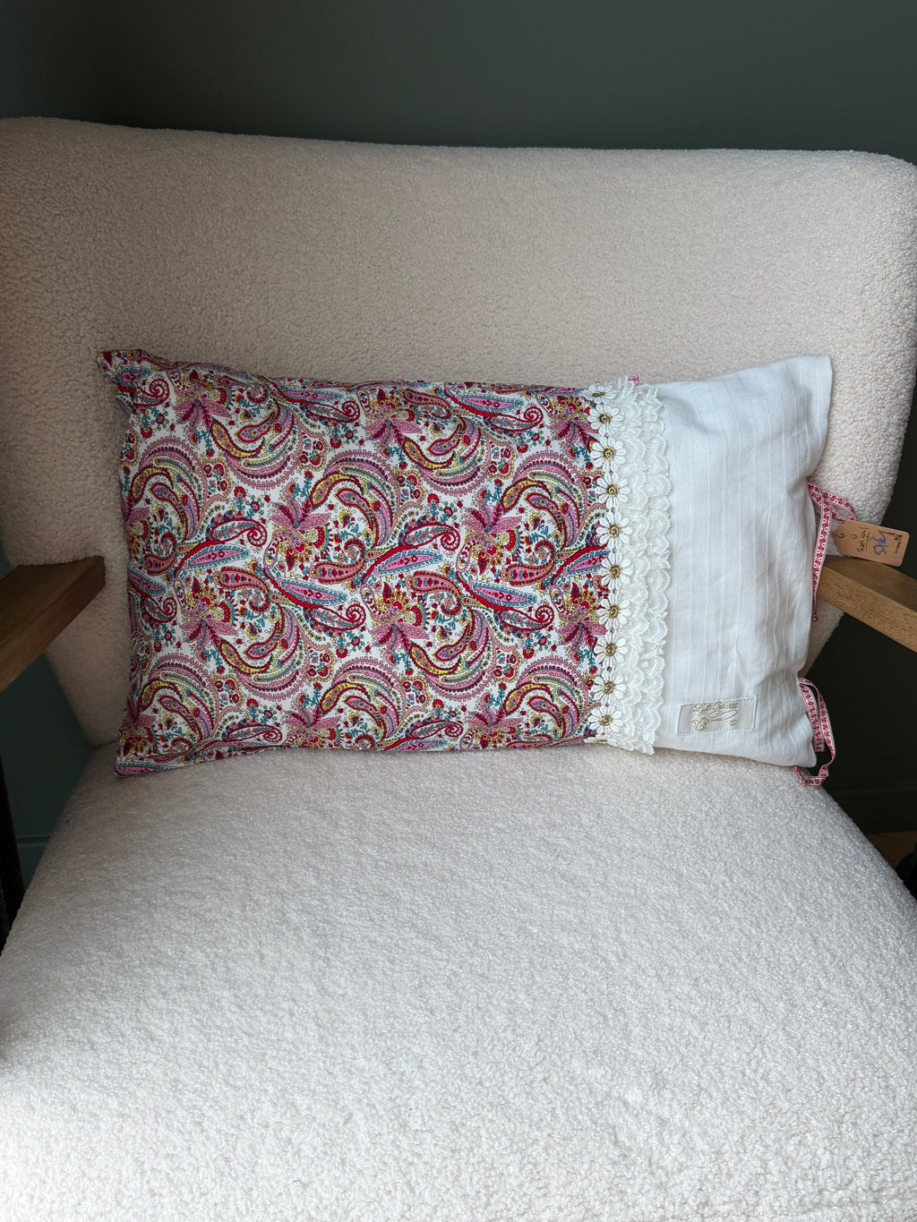 Coussin rectangulaire fait main, tissu imprimé cachemire rose et rouge et lin blanc rayé, dentelle fleurie et étiquette artisanale, posé sur fauteuil en bouclette blanche