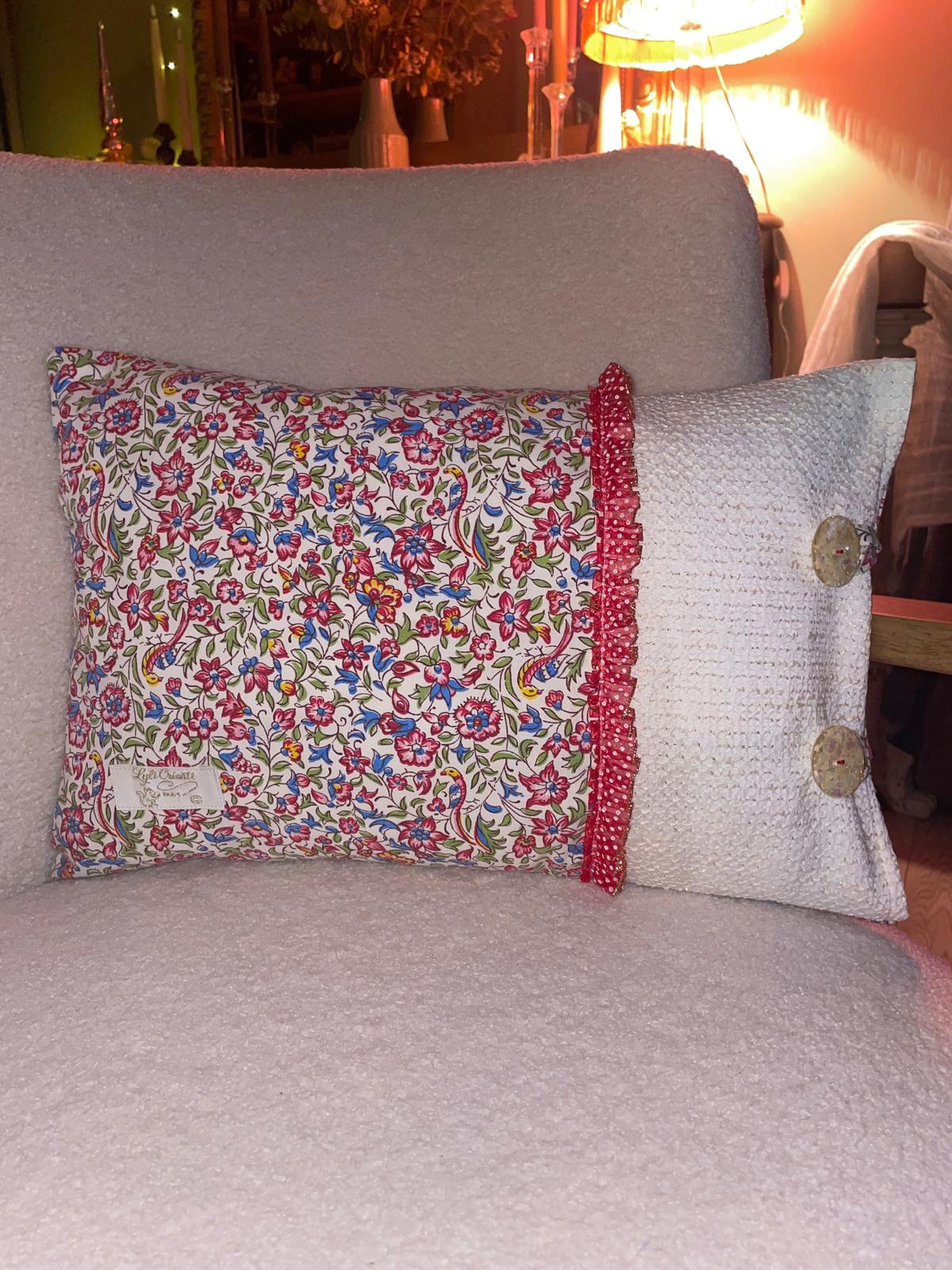 Coussin rectangulaire fait main Lyli Créarti, tissu imprimé fleuri oiseaux et lin blanc texturé, volant à pois rouge et gros boutons fantaisie, posé sur fauteuil en bouclette blanche