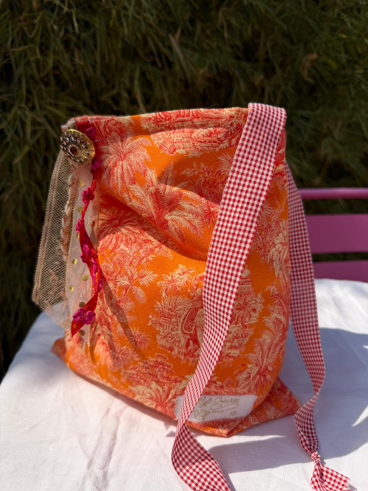 Bourse fait main en tissu imprimé toile de Jouy orange motif palmiers, anse longue à carreaux vichy rouge et blanc, posée sur table en extérieur