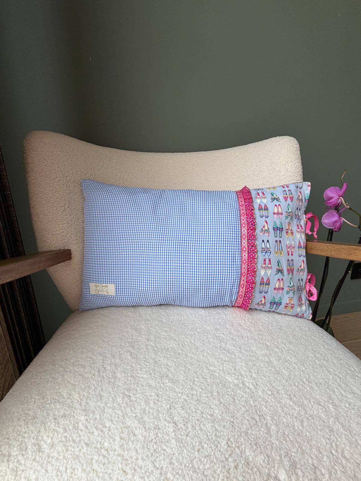 Coussin rectangulaire en coton à carreaux vichy bleu et blanc, bande imprimée motif chaussures et liseré rose, posé sur un fauteuil en bouclette blanche