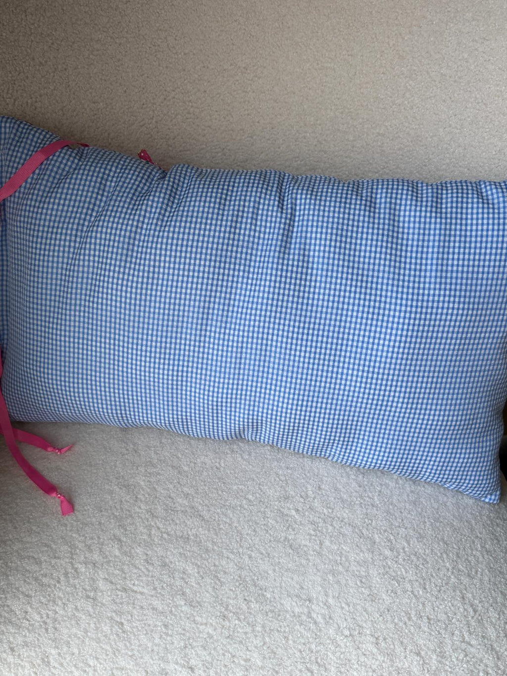 Coussin rectangulaire en coton vichy bleu et blanc avec rubans à pois rose, vue de face posé sur plaid en bouclette blanche
